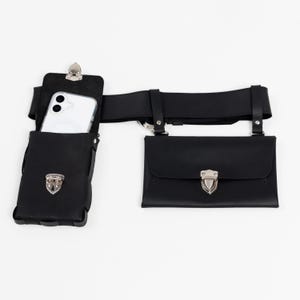 May include: Black leather belt bag with two pouches and a phone holder. The phone holder has a silver clip and holds a white smartphone. The pouches have silver clasps. The belt is black and has silver hardware.