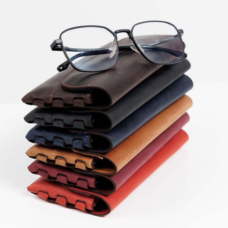 Pode incluir: Uma pilha de seis estojos de &oacute;culos de couro em v&aacute;rias cores: castanho, preto, azul marinho, bege, bord&ocirc; e vermelho. Um par de &oacute;culos com arma&ccedil;&atilde;o preta repousa no topo da pilha.