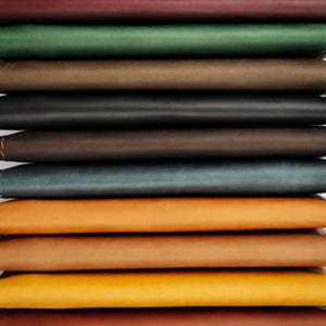 K&ouml;nnte beinhalten: Ein Stapel Leder-Notizb&uuml;cher in verschiedenen Farben, darunter Burgunderrot, Gr&uuml;n, Braun, Schwarz und T&uuml;rkis. Die Notizb&uuml;cher sind in einem ordentlichen Stapel angeordnet und zeigen die verschiedenen Farbt&ouml;ne und Texturen des Leders.