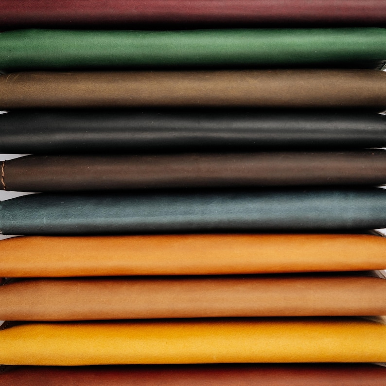 K&ouml;nnte beinhalten: Ein Stapel Leder-Notizb&uuml;cher in verschiedenen Farben, darunter Burgunderrot, Gr&uuml;n, Braun, Schwarz und T&uuml;rkis. Die Notizb&uuml;cher sind in einem ordentlichen Stapel angeordnet und zeigen die verschiedenen Farbt&ouml;ne und Texturen des Leders.
