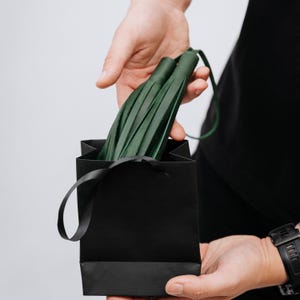 K&ouml;nnte beinhalten: Eine schwarze Geschenkt&uuml;te mit schwarzem Band wird gehalten. In der Tasche befindet sich ein gr&uuml;ner Lederflogger. Der Flogger besteht aus mehreren Streifen aus gr&uuml;nem Leder.