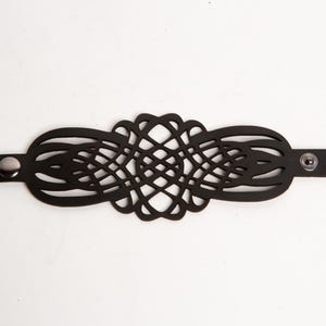 May include: Black leather bracelet with a detailed, openwork design. The bracelet has a central, symmetrical pattern and is fastened with snap closures. The bracelet is displayed on a white background.