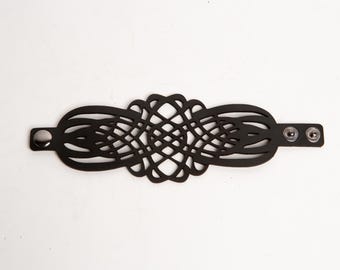 Puede incluir: Pulsera de cuero negro con un diseño intrincado y calado. La pulsera presenta un patrón central simétrico y se asegura con cierres de botón a presión. Se muestra sobre un fondo blanco.