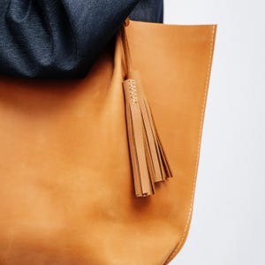 K&ouml;nnte beinhalten: Eine hellbraune Ledertasche mit passendem Quastenanh&auml;nger. Die Tasche hat eine hellbraune Farbe mit wei&szlig;en N&auml;hten entlang der Kanten. Die Quaste ist mit einem d&uuml;nnen Lederriemen an der Tasche befestigt.