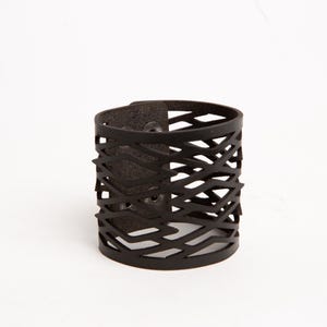 Puede incluir: Un brazalete de cuero marr&oacute;n oscuro con un dise&ntilde;o geom&eacute;trico recortado. El brazalete tiene un cierre de bot&oacute;n a presi&oacute;n y una forma cil&iacute;ndrica. El patr&oacute;n recortado crea un efecto tejido. El brazalete mide aproximadamente 5 cm de ancho.