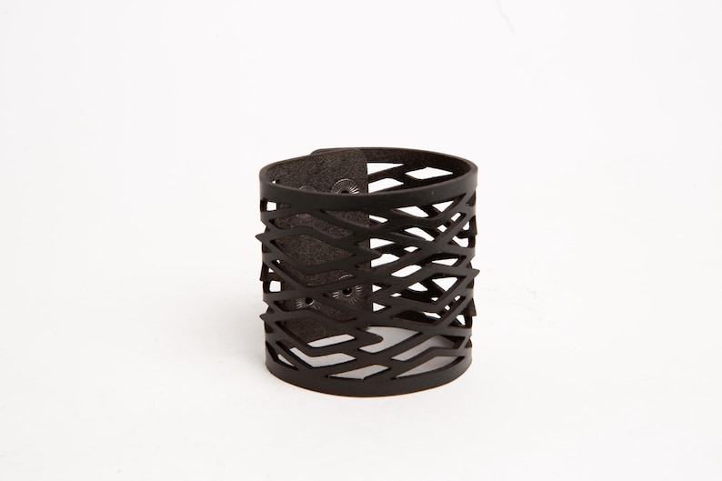 Puede incluir: Un brazalete de cuero marr&oacute;n oscuro con un dise&ntilde;o geom&eacute;trico recortado. El brazalete tiene un cierre de bot&oacute;n a presi&oacute;n y una forma cil&iacute;ndrica. El patr&oacute;n recortado crea un efecto tejido. El brazalete mide aproximadamente 5 cm de ancho.