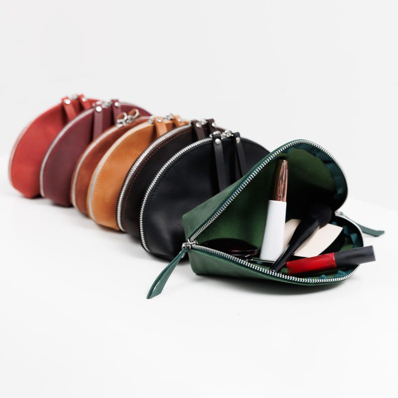 K&ouml;nnte beinhalten: Eine Sammlung halbmondf&ouml;rmiger Kosmetiktaschen in verschiedenen Farben, darunter Rot, Braun, Beige, Schwarz und Gr&uuml;n. Eine Tasche ist ge&ouml;ffnet und zeigt Make-up-Artikel. Jede Tasche hat einen silbernen Rei&szlig;verschluss.