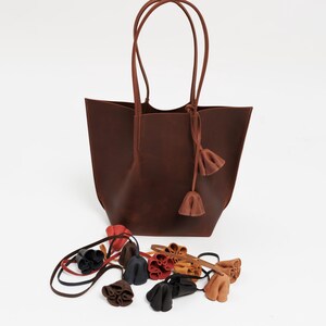 May include: A brown leather tote bag with long handles and decorative tassels. Several small, colorful leather accessories are scattered in front of the bag. The accessories include red, black, and brown pieces.
