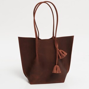 May include: A brown leather tote bag with long handles and a decorative tassel. The bag has a rectangular shape with a wide opening and is made of a smooth, solid material. The handles are attached at the top and are also brown.