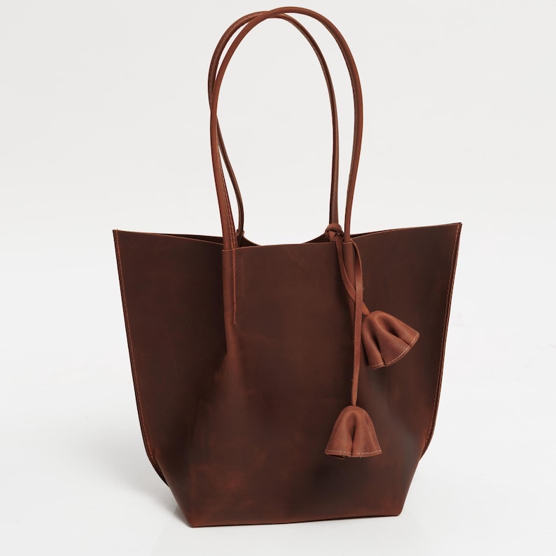 May include: A brown leather tote bag with long handles and a decorative tassel. The bag has a rectangular shape with a wide opening and is made of a smooth, solid material. The handles are attached at the top and are also brown.