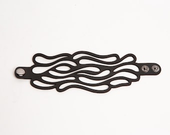 Puede incluir: Pulsera de cuero negro con un diseño calado único y fluido. La pulsera presenta una serie de formas onduladas interconectadas. Se abrocha con dos cierres de presión y un solo botón de presión. La pulsera mide aproximadamente 3,8 cm de ancho.