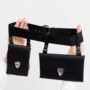 May include: Black leather belt with two attached pouches. The belt has a silver buckle and silver clasps on the pouches. The pouches have silver clasps. The belt is held up by a person's hands.