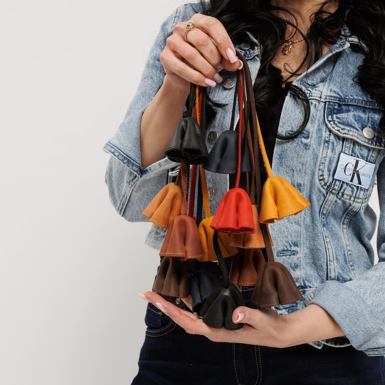 May include: A collection of leather key fobs in various colors, including black, brown, orange, and yellow. Each fob has a bell-shaped design and is attached to a thin leather strap. The key fobs are held up against a light background.