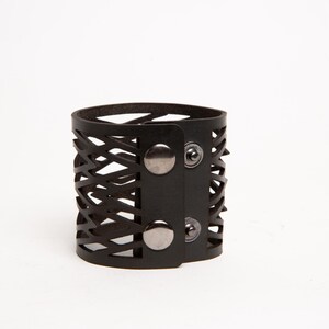 Puede incluir: Un brazalete de cuero negro con un dise&ntilde;o geom&eacute;trico recortado. El brazalete presenta un estilo envolvente con cierre de bot&oacute;n a presi&oacute;n. El brazalete mide aproximadamente 5 cm de ancho y tiene una est&eacute;tica moderna y atrevida.