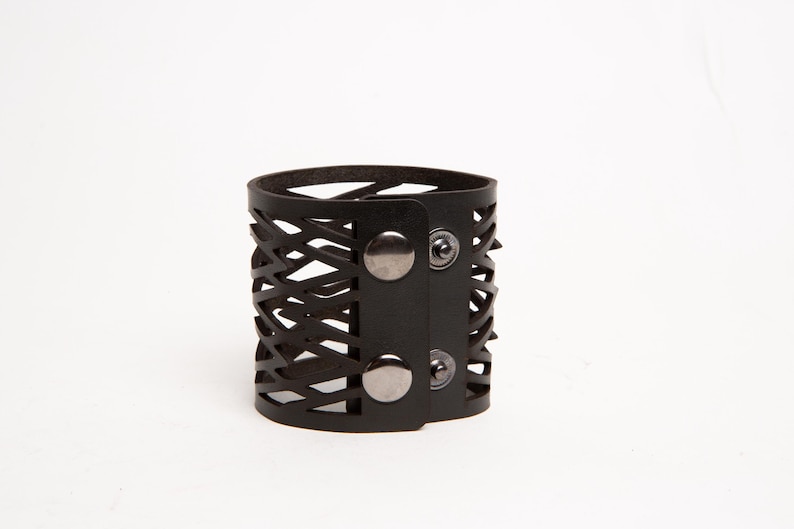Puede incluir: Un brazalete de cuero negro con un dise&ntilde;o geom&eacute;trico recortado. El brazalete presenta un estilo envolvente con cierre de bot&oacute;n a presi&oacute;n. El brazalete mide aproximadamente 5 cm de ancho y tiene una est&eacute;tica moderna y atrevida.
