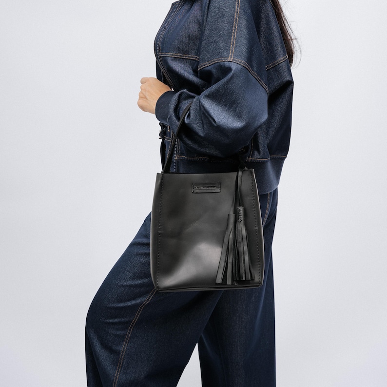 K&ouml;nnte beinhalten: Schwarze Leder-Eimertasche mit langem Schulterriemen und Quaste. Die Tasche hat eine rechteckige Form und wird von einer Person getragen, die eine Jeansjacke und Jeans tr&auml;gt.