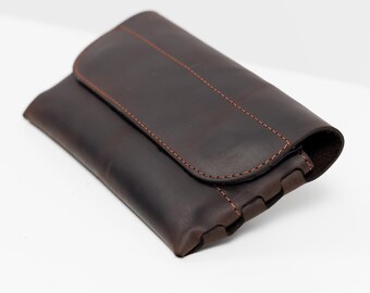 May include: A dark brown leather clutch with a flap closure. The clutch features orange stitching along the edges and a textured surface. The folded design creates a compact shape, suitable for carrying essentials.