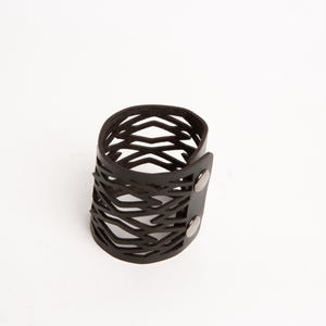 Puede incluir: Una pulsera de cuero negro con un dise&ntilde;o geom&eacute;trico calado. La pulsera es cil&iacute;ndrica y se abrocha con dos broches plateados. El patr&oacute;n calado crea una est&eacute;tica &uacute;nica y moderna. Este accesorio es adecuado para el uso diario.