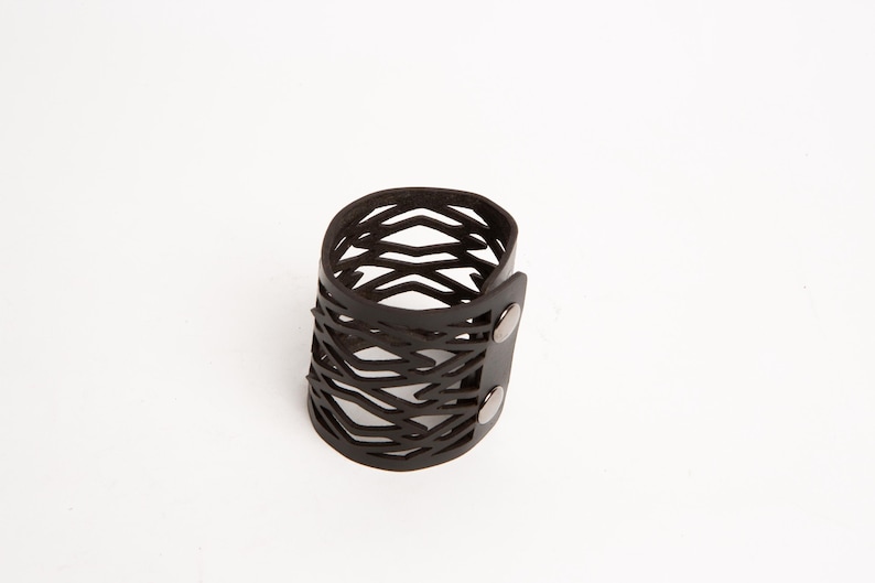 Puede incluir: Una pulsera de cuero negro con un dise&ntilde;o geom&eacute;trico calado. La pulsera es cil&iacute;ndrica y se abrocha con dos broches plateados. El patr&oacute;n calado crea una est&eacute;tica &uacute;nica y moderna. Este accesorio es adecuado para el uso diario.