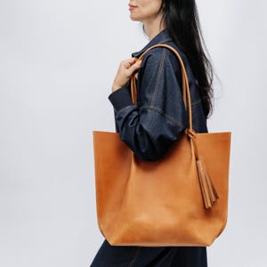 K&ouml;nnte beinhalten: Eine gro&szlig;e, hellbraune Ledertasche mit langen Henkeln und einem dekorativen Quastenanh&auml;nger. Die Tasche wird &uuml;ber der Schulter getragen. Das Leder hat eine glatte Oberfl&auml;che und eine nat&uuml;rliche Farbe. Die Tasche ist rechteckig.
