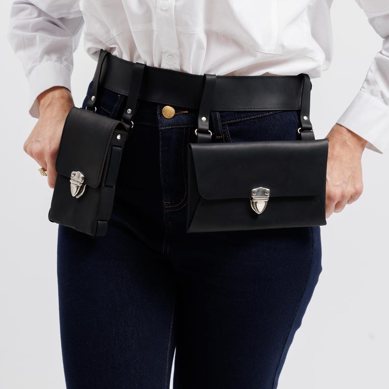 May include: Black leather belt bag with two pouches. Each pouch has a silver-colored clasp. The belt is worn with dark blue jeans and a white button-down shirt. The pouches are rectangular and have a flap closure.