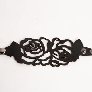 May include: Black leather cuff bracelet featuring a floral rose design. The bracelet has a cut-out pattern of roses and leaves. It fastens with two snap closures and a single silver-tone stud.