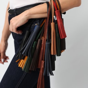 Puede incluir: Una colección de borlas de cuero en varios colores, incluyendo negro, azul marino, verde oscuro, marrón, tostado y rojo. Cada borla cuenta con una correa larga y está construida con múltiples tiras de cuero unidas con costuras visibles.