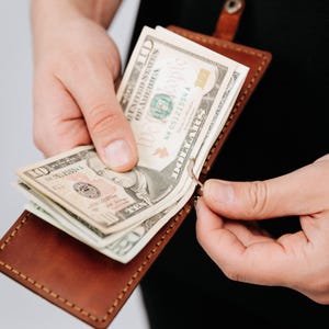 May include: A brown leather money clip holds several ten-dollar bills. The clip is open, revealing the bills, and is held by a person. The leather has a stitched border and a snap closure.