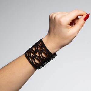 Puede incluir: Un brazalete negro con un dise&ntilde;o geom&eacute;trico calado. El brazalete es ancho y se envuelve alrededor de la mu&ntilde;eca. El dise&ntilde;o presenta una serie de formas interconectadas, creando una apariencia texturizada. El brazalete est&aacute; hecho de un material oscuro, posiblemente cuero o una sustancia similar.