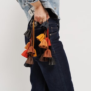 May include: A collection of leather bell-shaped charms in various colors, including black, brown, orange, and yellow, are held against a person's dark blue jeans. The charms are attached to thin leather cords.