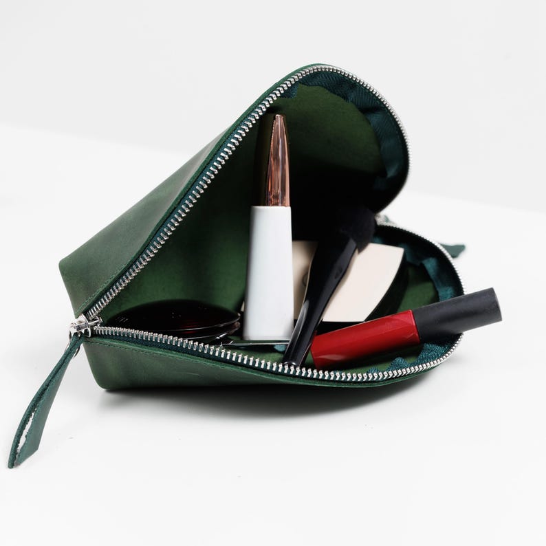 K&ouml;nnte beinhalten: Offene gr&uuml;ne Kosmetiktasche aus Leder mit silbernem Rei&szlig;verschluss, gef&uuml;llt mit Make-up-Artikeln wie Lippenstift, Pinsel und Kompaktpuder. Die Tasche ist ge&ouml;ffnet und zeigt das gr&uuml;ne Innere.
