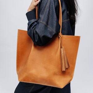 K&ouml;nnte beinhalten: Eine gro&szlig;e, hellbraune Ledertasche mit rechteckiger Form und Quaste. Die Tasche hat ein schlichtes Design mit sichtbaren N&auml;hten und langen Henkeln. Das Leder hat ein nat&uuml;rliches, leicht abgenutztes Aussehen.
