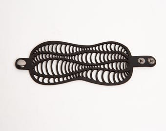 Puede incluir: Una pulsera de cuero negro con un distintivo diseño calado. El diseño presenta numerosos cortes verticales y curvos. Se abrocha con dos cierres de presión y un solo lazo. La forma de la pulsera es alargada y simétrica, de unos 20 cm de largo.