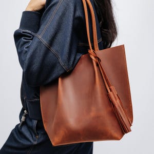 K&ouml;nnte beinhalten: Eine braune Ledertasche mit langen Riemen und einem Quastenanh&auml;nger. Die Tasche hat eine satte, warme braune Farbe und scheint aus hochwertigem Leder gefertigt zu sein. Die Riemen sind in einem helleren Braunton gehalten. Die Tasche wird &uuml;ber der Schulter getragen.