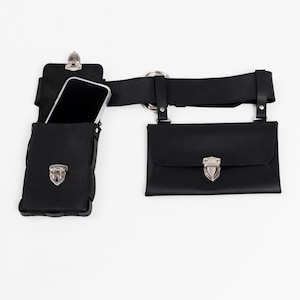 May include: Black leather belt with two pouches and a phone holder. The belt has a silver buckle and hardware. One pouch has a flap closure with a silver clasp. The phone holder is open at the top, and a phone is visible inside.