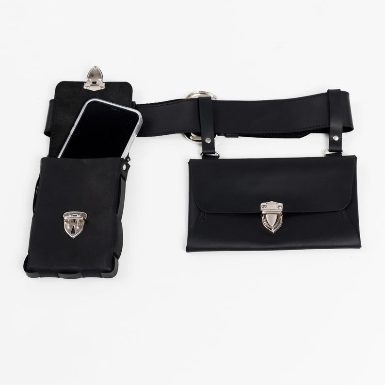 May include: Black leather belt with two pouches and a phone holder. The belt has a silver buckle and hardware. One pouch has a flap closure with a silver clasp. The phone holder is open at the top, and a phone is visible inside.