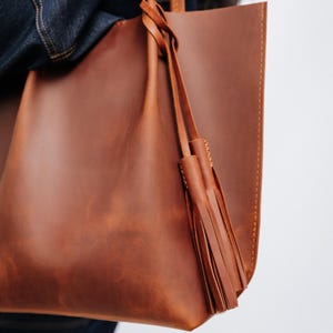 K&ouml;nnte beinhalten: Eine braune Ledertasche mit Quasten-Detail. Die Tasche besteht aus glattem, strukturiertem Leder und hat ein schlichtes, minimalistisches Design. Die Tasche wird von einer Person getragen, die eine Jeansjacke tr&auml;gt.