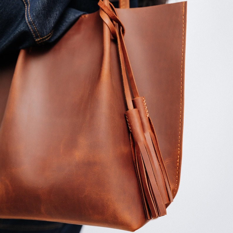 K&ouml;nnte beinhalten: Eine braune Ledertasche mit Quasten-Detail. Die Tasche besteht aus glattem, strukturiertem Leder und hat ein schlichtes, minimalistisches Design. Die Tasche wird von einer Person getragen, die eine Jeansjacke tr&auml;gt.