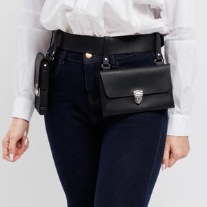 May include: Black leather belt bag with two pouches. The belt is worn around the waist and has a gold-colored buckle. The pouches have silver-colored clasps. The person in the image is wearing a white shirt and dark blue jeans.