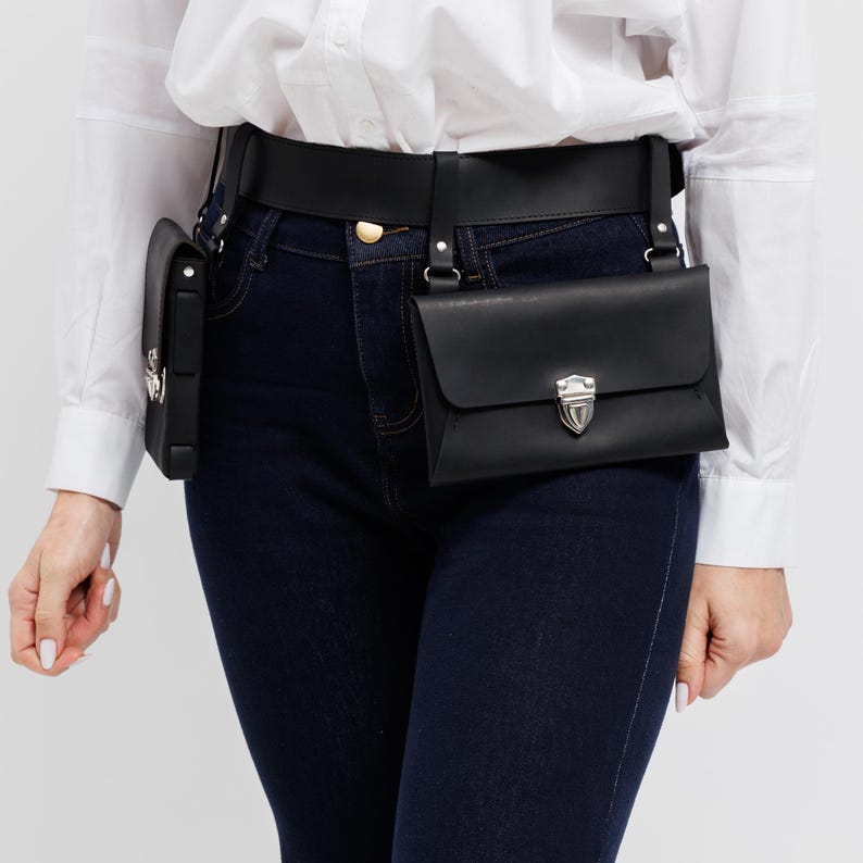 May include: Black leather belt bag with two pouches. The belt is worn around the waist and has a gold-colored buckle. The pouches have silver-colored clasps. The person in the image is wearing a white shirt and dark blue jeans.