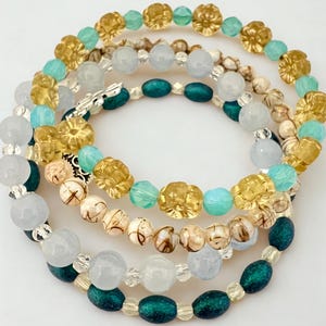 May include: Four beaded bracelets in shades of gold, teal, white, and brown. The bracelets feature various bead shapes, including round, flower-shaped, and oval beads. The bracelets are stacked on a white surface.