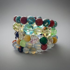 May include: A stack of colorful beaded bracelets. The bracelets feature a variety of beads in shades of red, turquoise, green, yellow, and clear. The beads are round and appear to be made of glass or a similar material. The bracelets are stacked together, creating a layered effect.