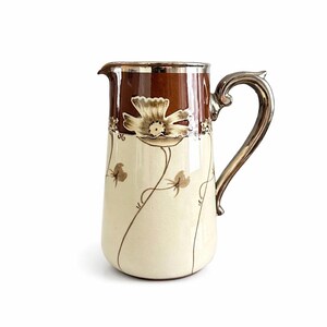 Peut inclure: Pichet crème avec un dessus marron et une poignée argentée. Le pichet présente un motif floral avec une grande fleur et des tiges délicates. La poignée et le bord sont argentés, ajoutant une touche d'élégance au pichet de style vintage.