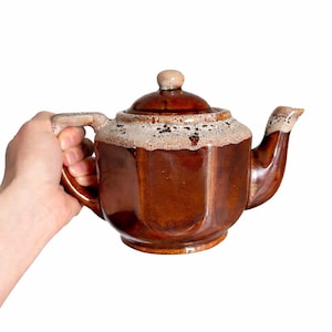May include: A brown ceramic teapot with a lid and spout. The teapot has a geometric design and a light-coloured rim. The handle is held by a person. A classic piece for serving tea.