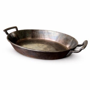 May include: Oval-shaped, antique-style metal pan with two curved handles. The pan has a dark, aged appearance, with visible wear and tear. The interior has a lighter, reflective surface. The pan is designed for cooking and serving.