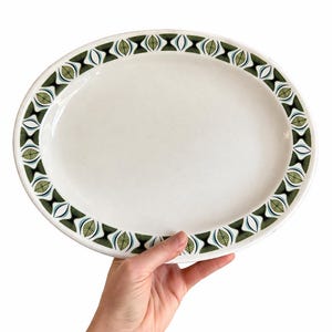 May include: Oval serving platter with a white surface and a decorative border. The border features a repeating pattern of green, black, and blue leaf-like shapes. The platter is held by a hand against a white background.