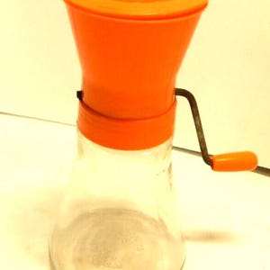 Op de afbeelding: Een oranje en heldere glazen voedselmolen met een slinger. De molen heeft een conische glazen basis en een oranje plastic bovenkant met een handvat. De slinger is van metaal met een oranje plastic handvat.