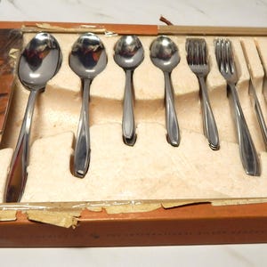 Könnte beinhalten: Ein Vintage-Set aus Edelstahl-Besteck, einschließlich Löffel, Gabeln und Messer, präsentiert in einer Pappschachtel. Die Schachtel hat ein braunes und orangefarbenes Farbschema mit dem Text "Stainless Steel Tableware by INTERNATIONAL".
