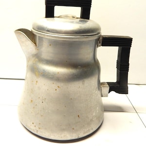 Puede incluir: Una cafetera vintage de aluminio con asa y asa de tapa negras. La cafetera tiene un pico y un pomo de vidrio transparente en la tapa. El exterior muestra signos de desgaste.