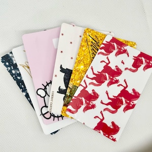 Pu&ograve; includere: Una collezione di carte rettangolari con vari disegni. Una carta ha un motivo a stelle in bianco e nero, un'altra presenta un design floreale dorato e una carta rosa ha una faccia da cartone animato. Altre carte hanno grafiche a tema cavallo e cowboy.